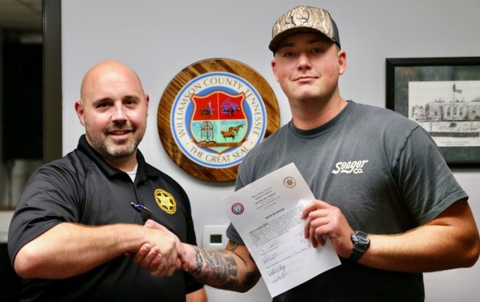 Capt. Chad Youker, left, shakes hands with newly sworn-in Detention Deputy Blake Larson, right, who holds his oath of office. The Williamson County, Tennessee, seal is displayed on the wall behind them.