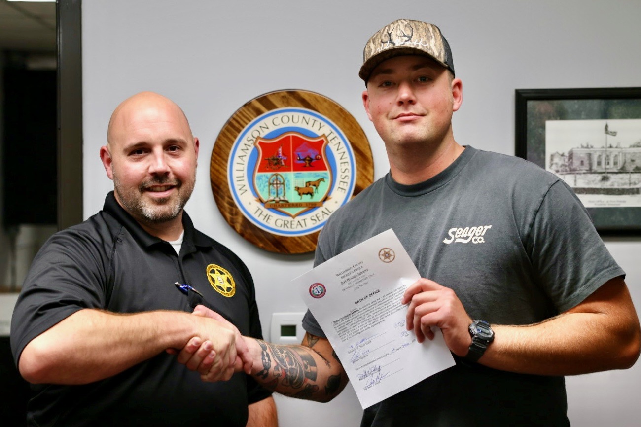 Capt. Chad Youker, left, shakes hands with newly sworn-in Detention Deputy Blake Larson, right, who holds his oath of office. The Williamson County, Tennessee, seal is displayed on the wall behind them.