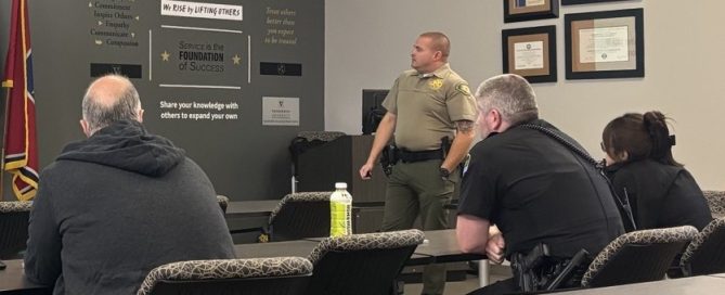 Law enforcement officers sit at classroom tables listening to a DUI enforcement training presentation, with several officers taking notes and facing the instructor.