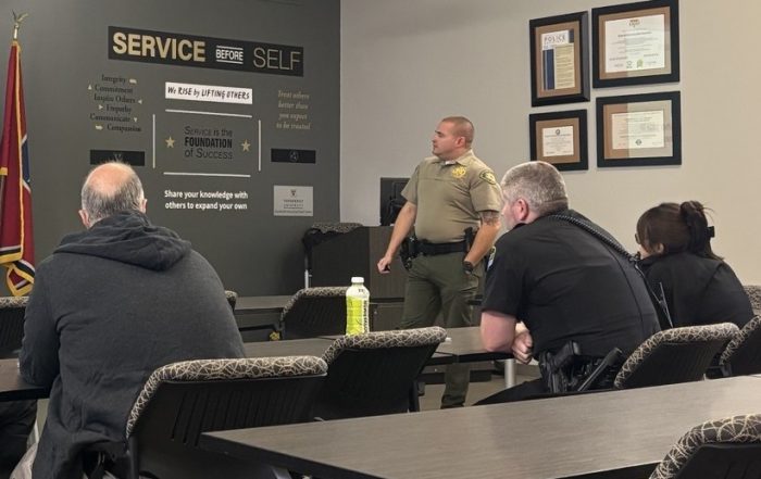 Law enforcement officers sit at classroom tables listening to a DUI enforcement training presentation, with several officers taking notes and facing the instructor.