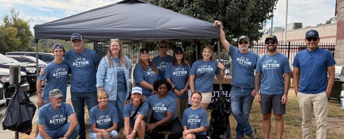 A group of Southall Church volunteers pose together outdoors beneath a canopy during a Deputy Appreciation Cookout. Many are wearing matching blue “Love in Action” shirts, with food and grilling equipment set up behind them.