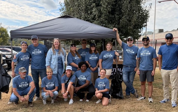 A group of Southall Church volunteers pose together outdoors beneath a canopy during a Deputy Appreciation Cookout. Many are wearing matching blue “Love in Action” shirts, with food and grilling equipment set up behind them.